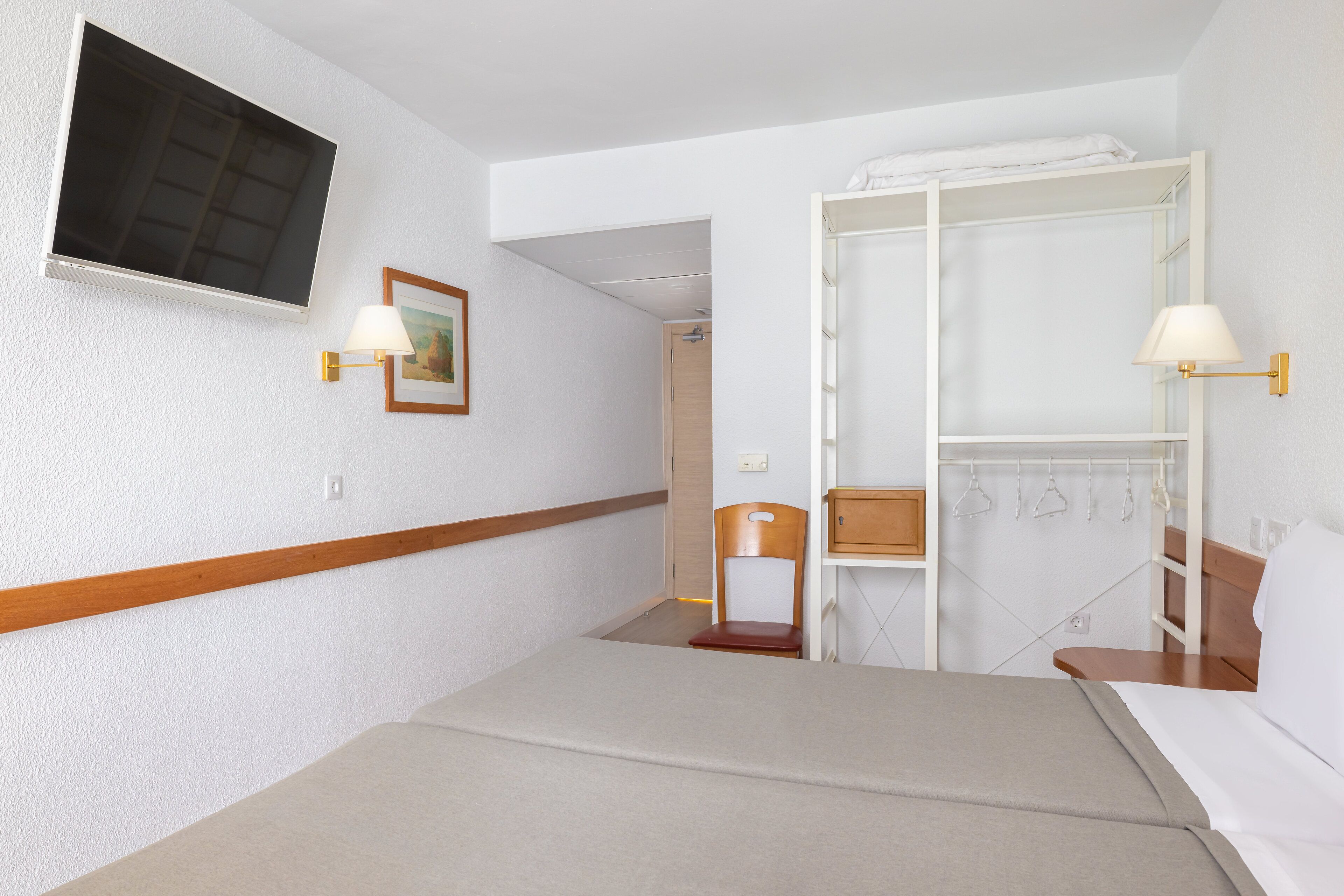 A hotel room with a bed, a TV mounted on the wall, a wooden chair, a desk with a lamp, and a wardrobe with hanging clothes.