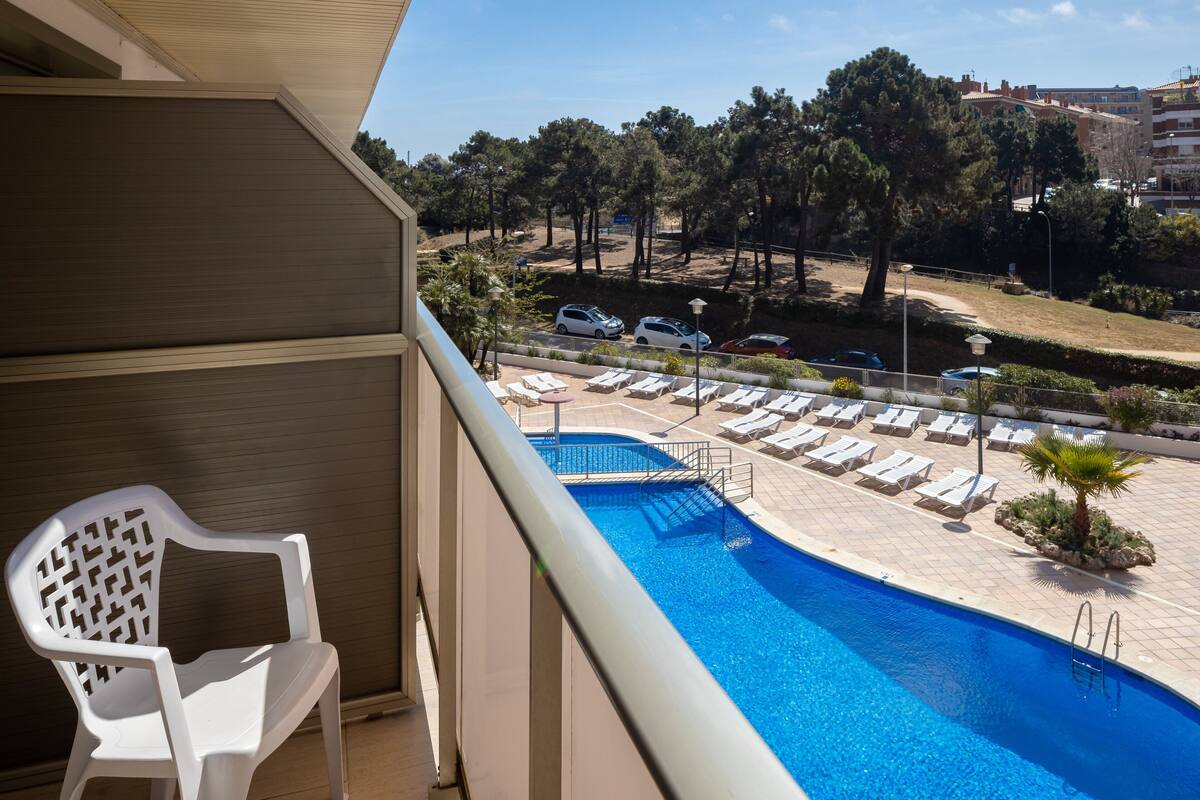 Un balcón con vista a una piscina, sillones de descanso y un estacionamiento con coches.