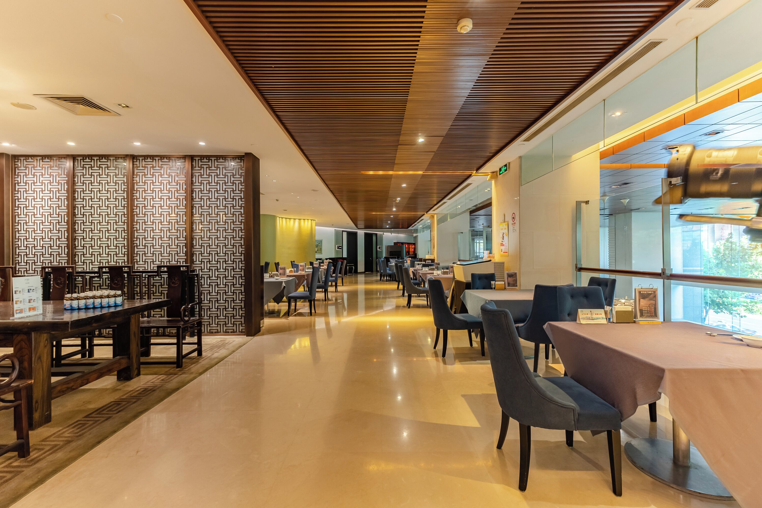 A modern restaurant with a long wooden table and chairs, a patterned wall, and a view through large windows.