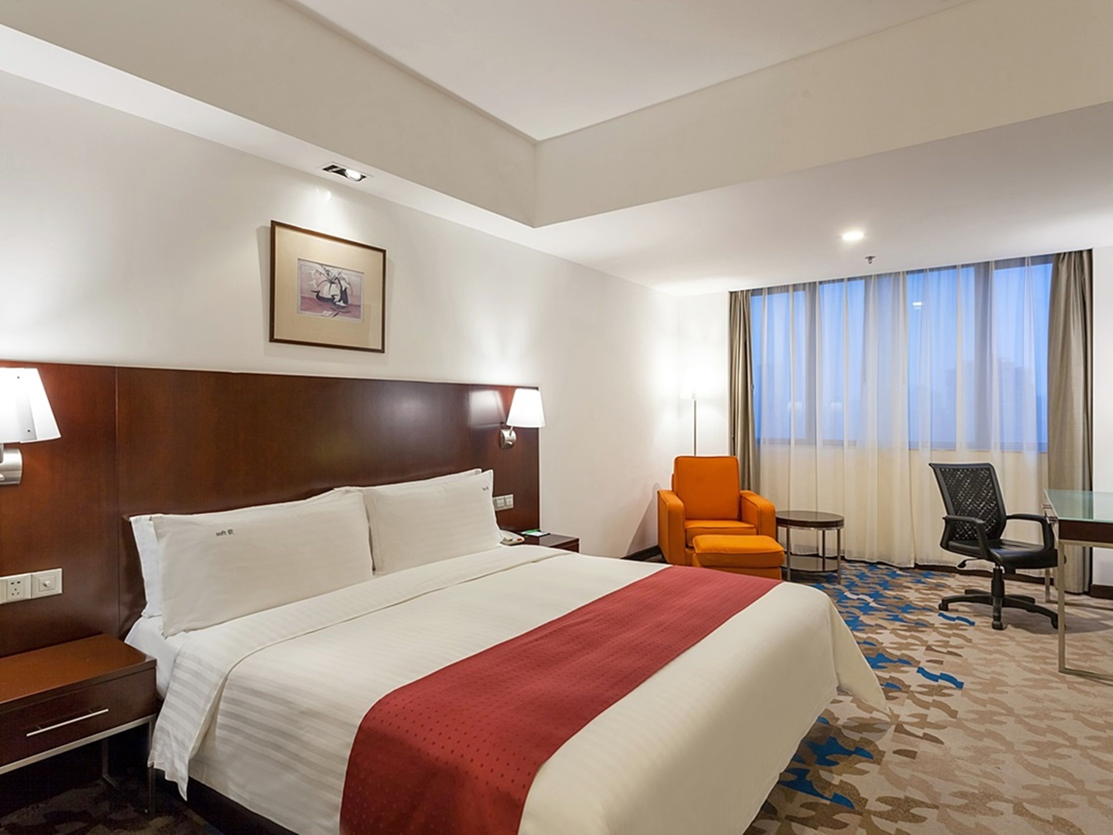 A hotel room with a large bed, a desk, an orange chair, and a view of the outdoors through the window.
