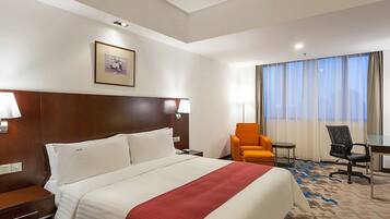 A hotel room with a large bed, a desk, an orange chair, and a view of the outdoors through the window.