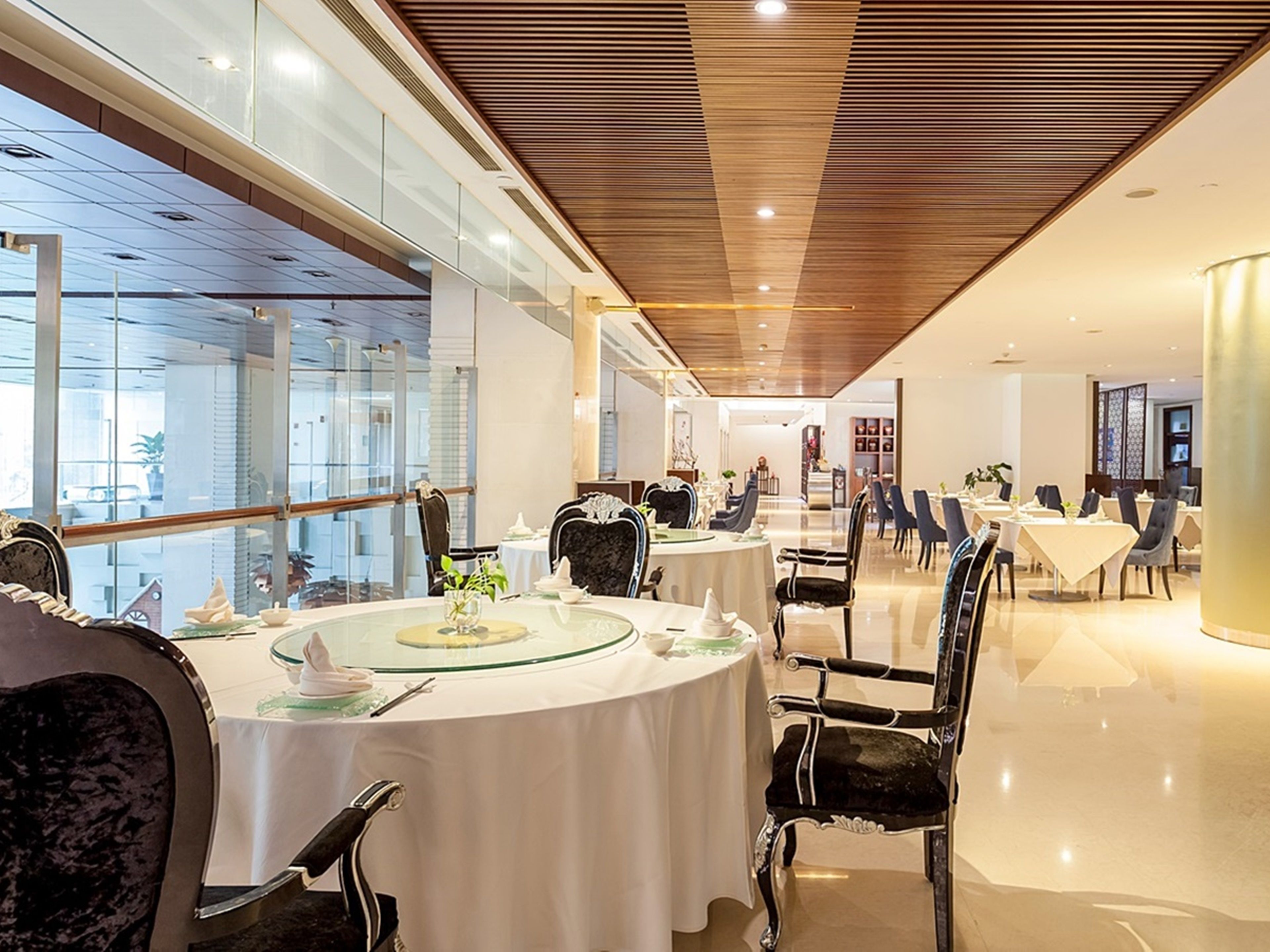 A modern dining area with round tables and black upholstered chairs, large windows, and a wooden ceiling.