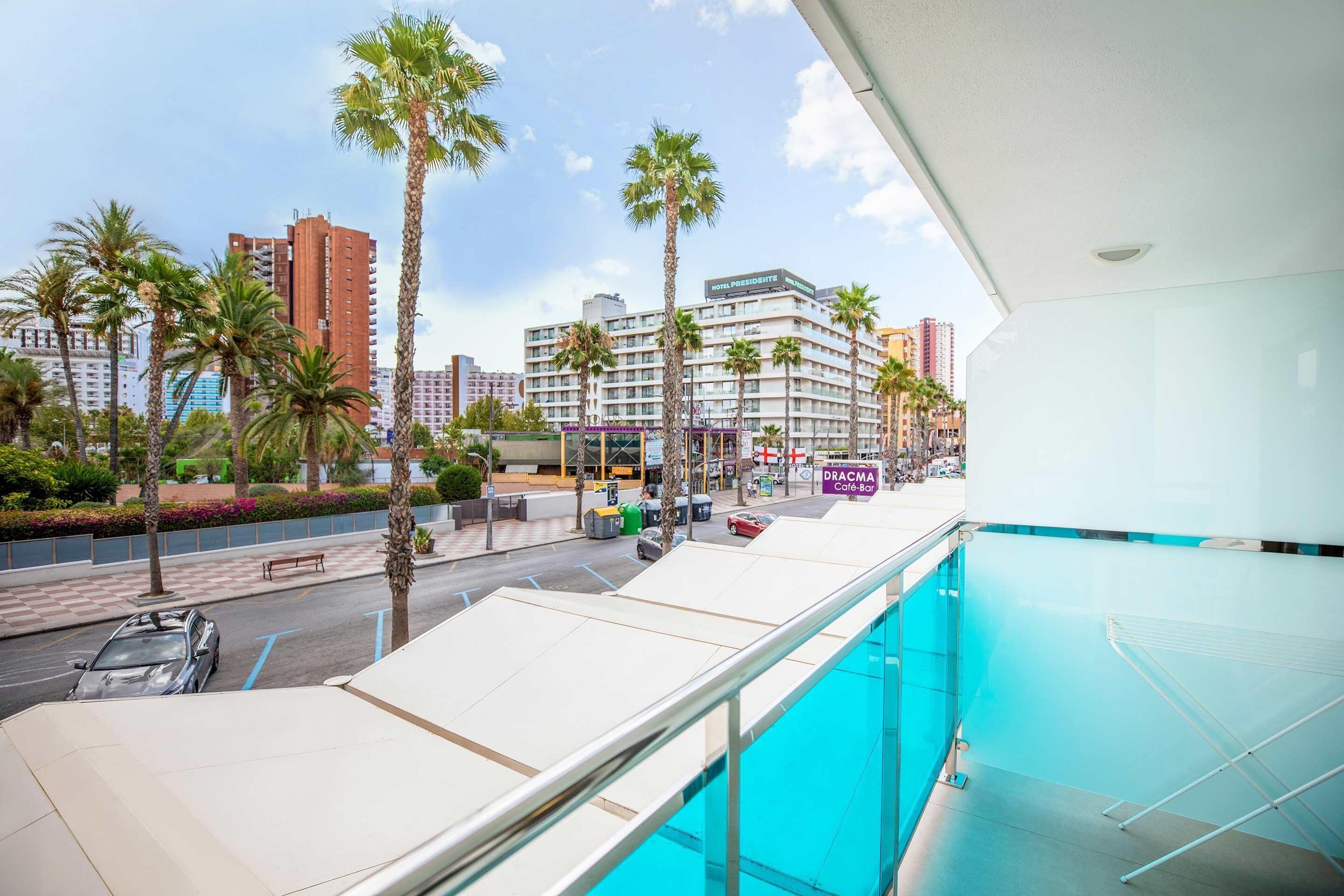 A hotel balcony with a pool, palm trees, and a view of the street and buildings.