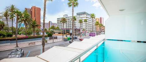 A hotel balcony with a pool, palm trees, and a view of the street and buildings.