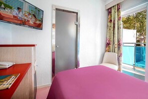 Quarto de hotel com cama, escrivaninha com telefone e revista, porta e varanda com cadeira.