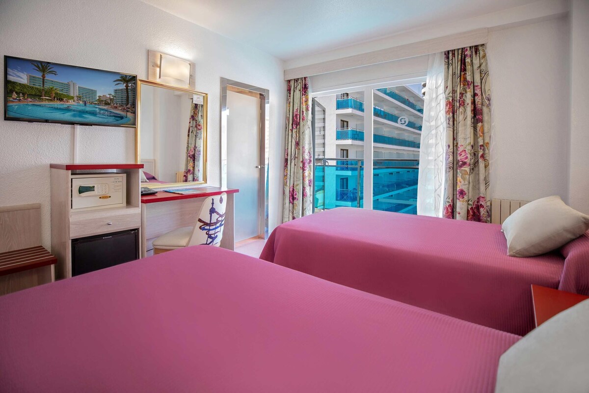 A hotel room with two beds, a desk, a small fridge, and a balcony with a pool view.