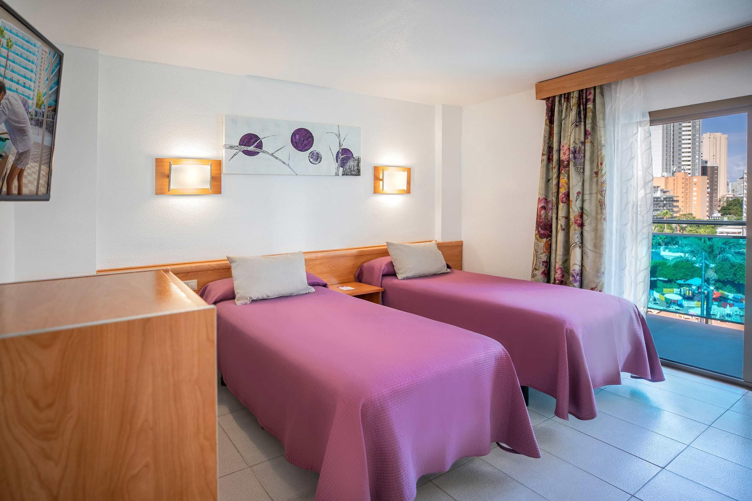 A hotel room with two beds, a wooden desk, a city view balcony, and wall-mounted lights.