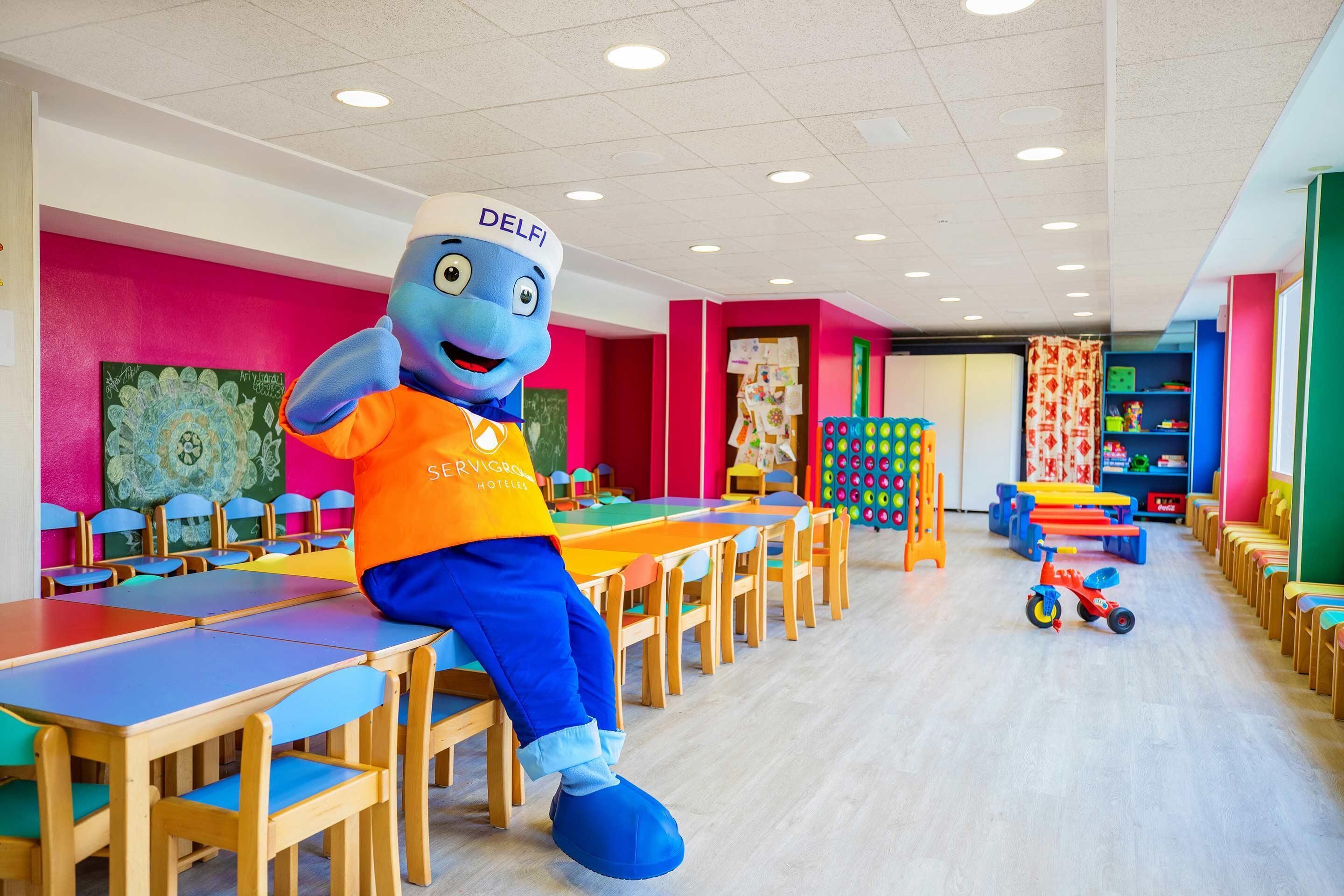 Un personaje promocional en una habitación colorida con mesas y sillas, una mesita con juguetes, y una pared con un diseño llamativo.
