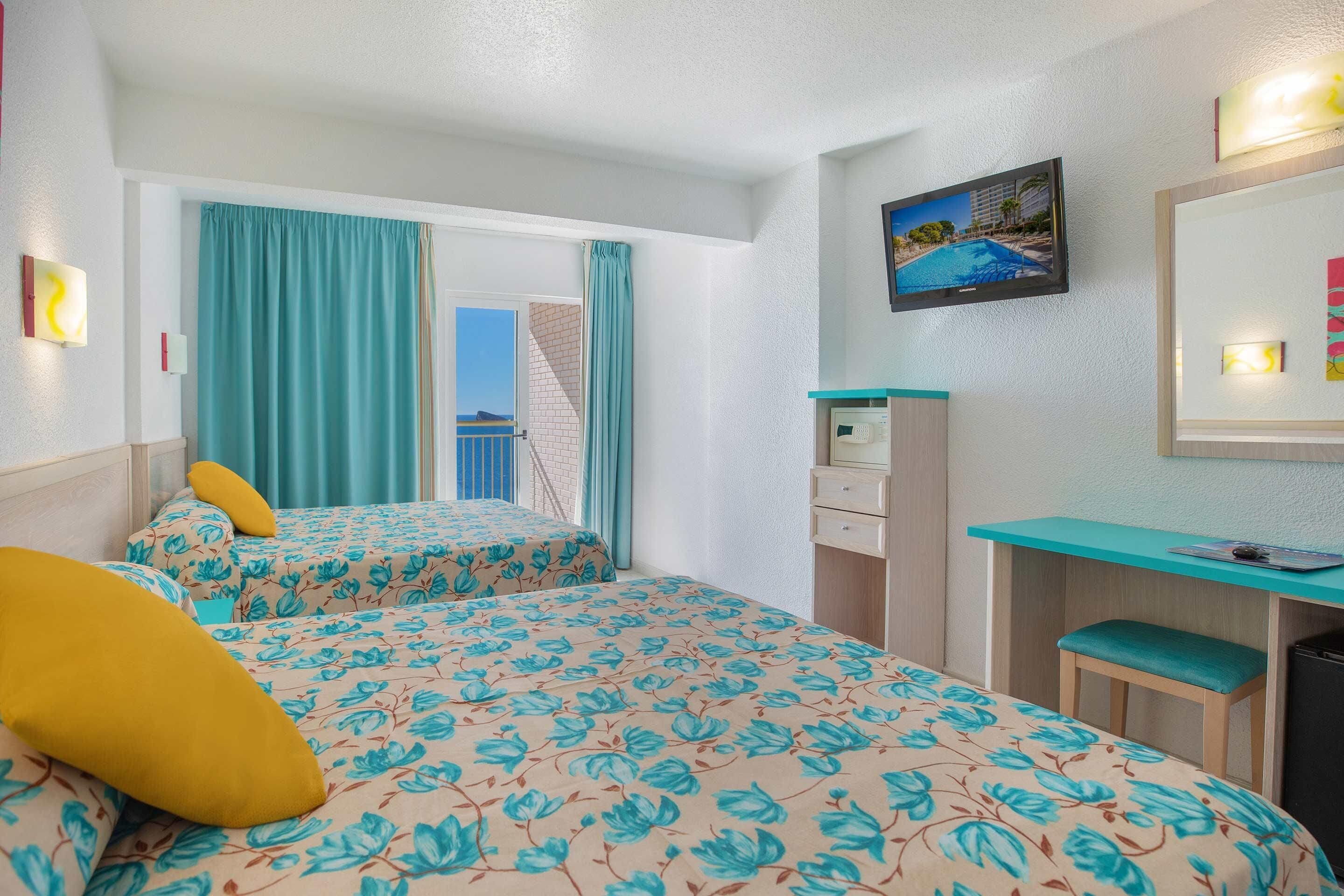 Una camera d'albergo con due letti, una scrivania, una sedia, una televisione e una finestra con tende.
