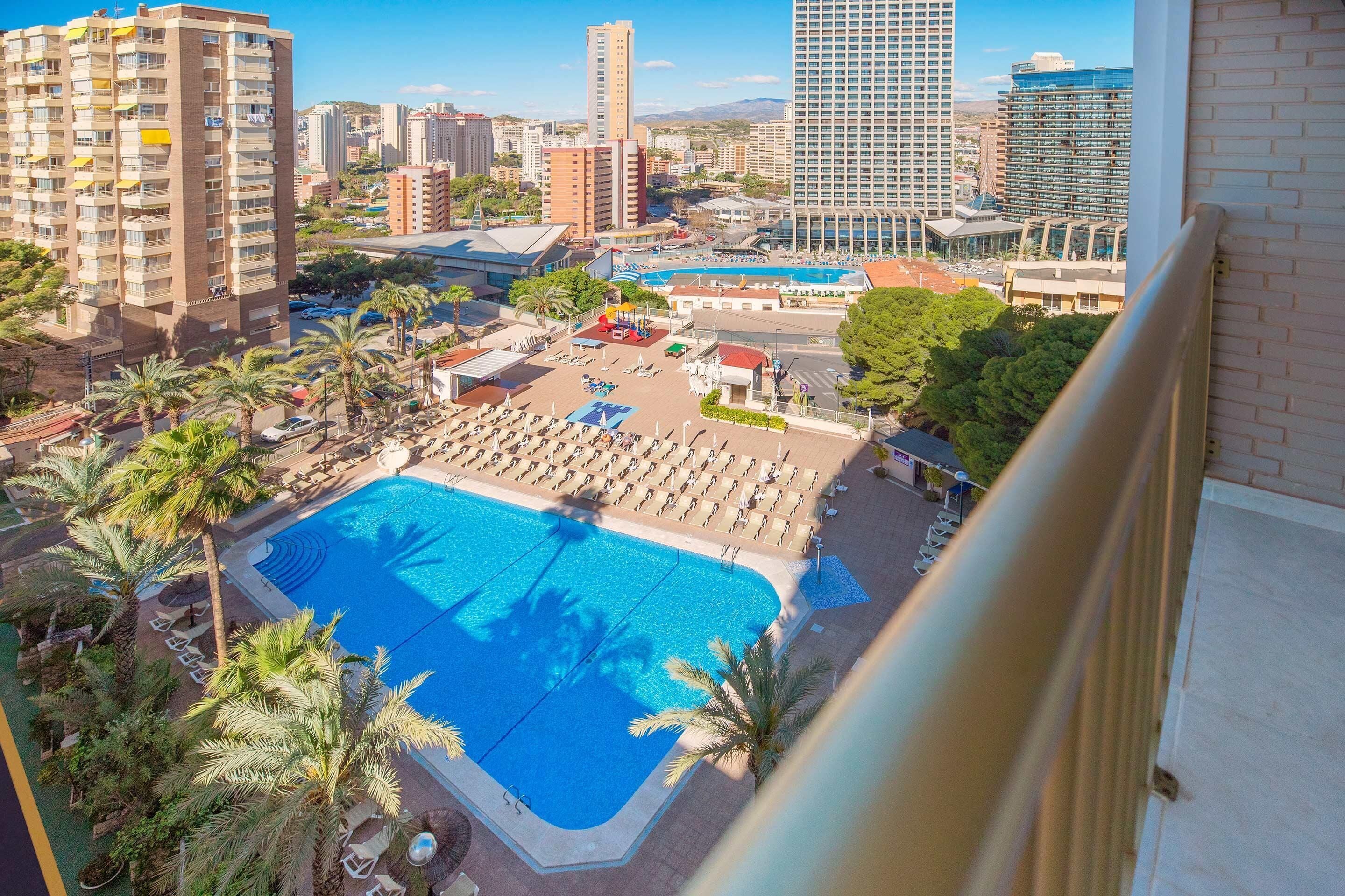 A hotel with a large swimming pool surrounded by sun loungers and palm trees.