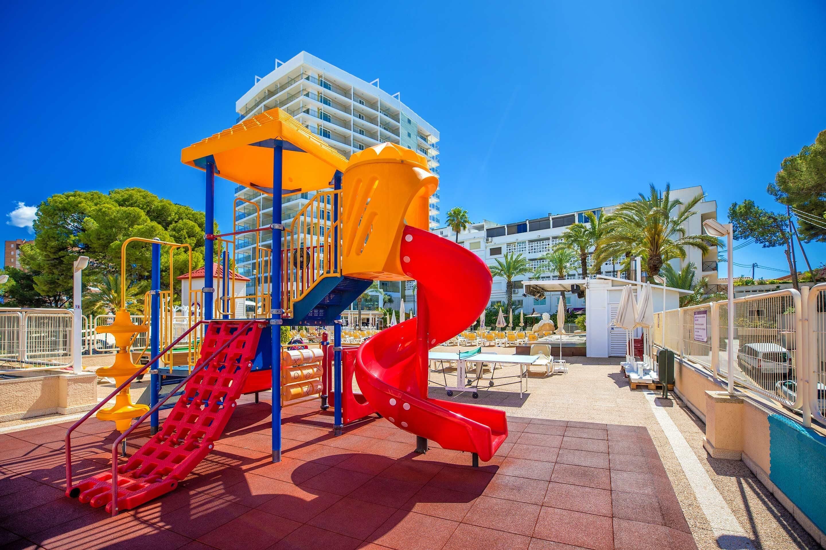A playground with a red spiral slide and yellow climbing structures, surrounded by a tiled area and palm trees.