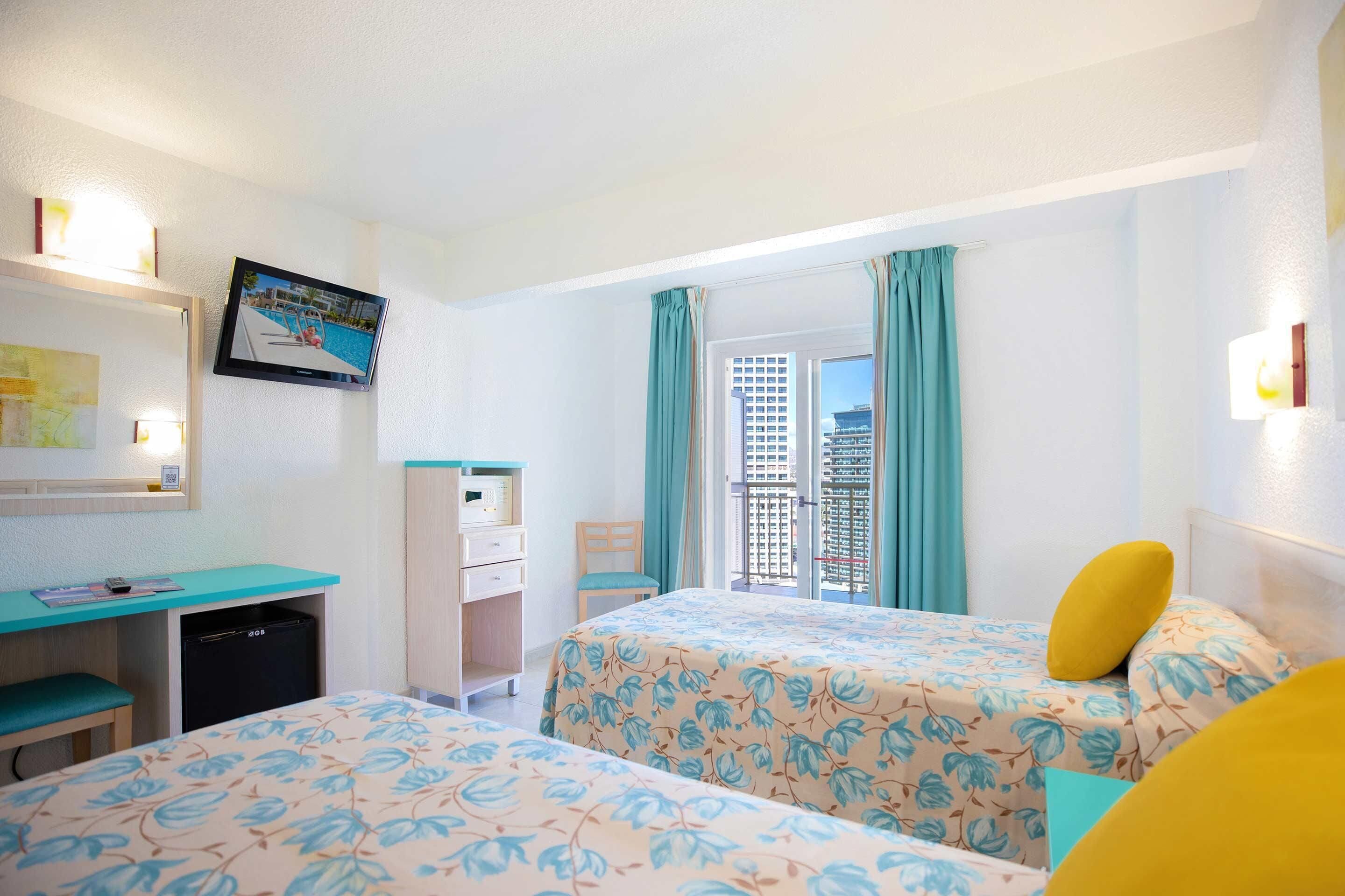 A hotel room with two beds, a desk, a small fridge, a window with curtains, and a TV on the wall.