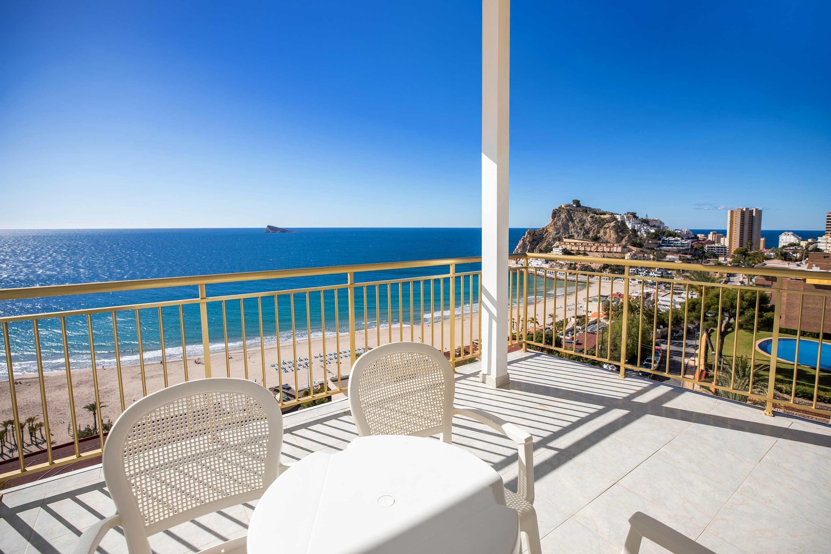 A balcony with white chairs and a table, overlooking a beach and a coastal town.
