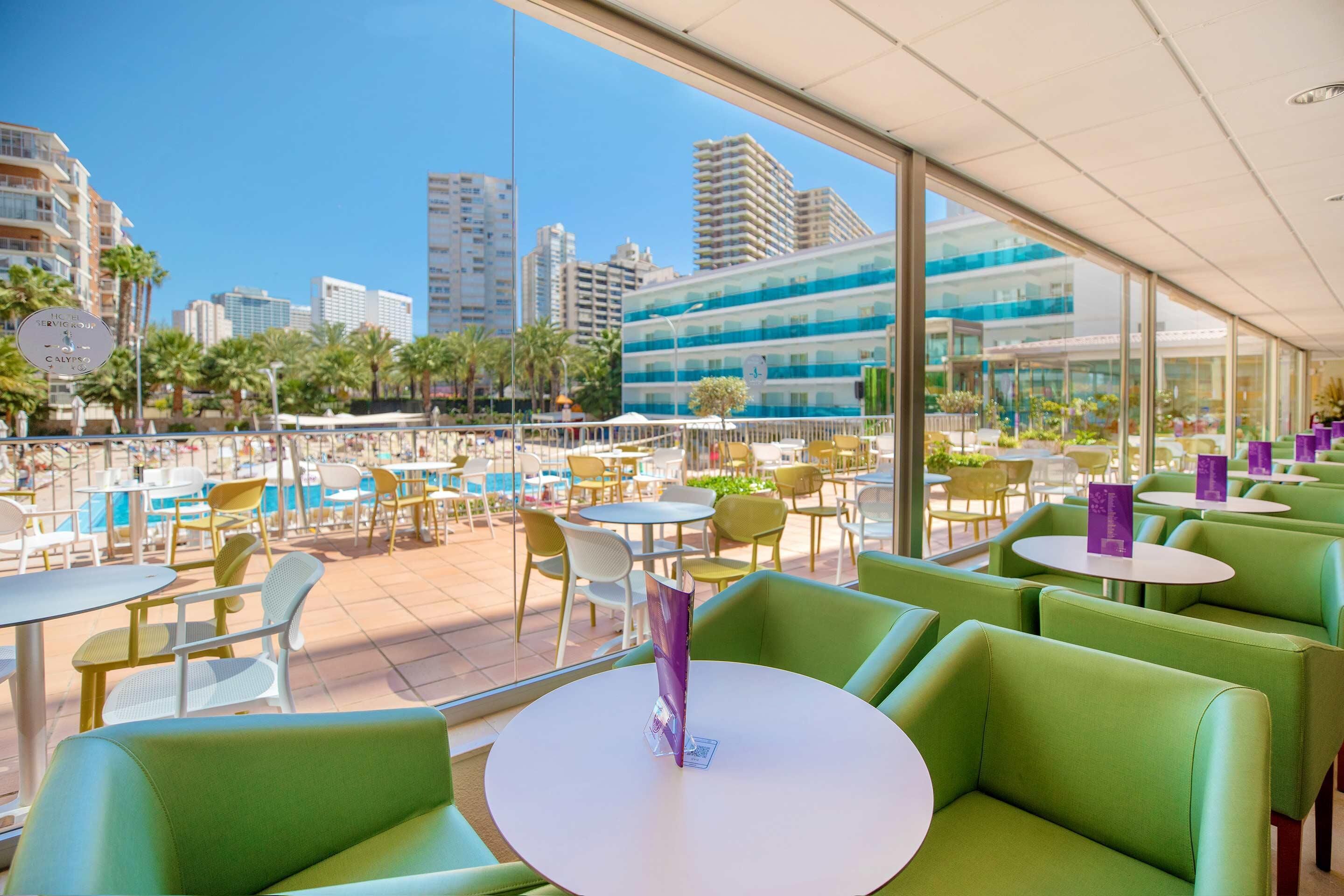 Um bar no terraço com assentos verdes, piscina e vista para a cidade.