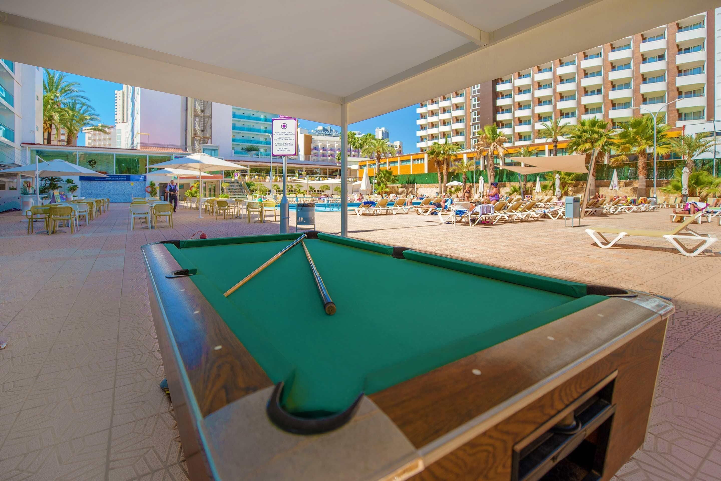 Una mesa de billar en el vestíbulo de un hotel, con zona de piscina y sillones de descanso.
