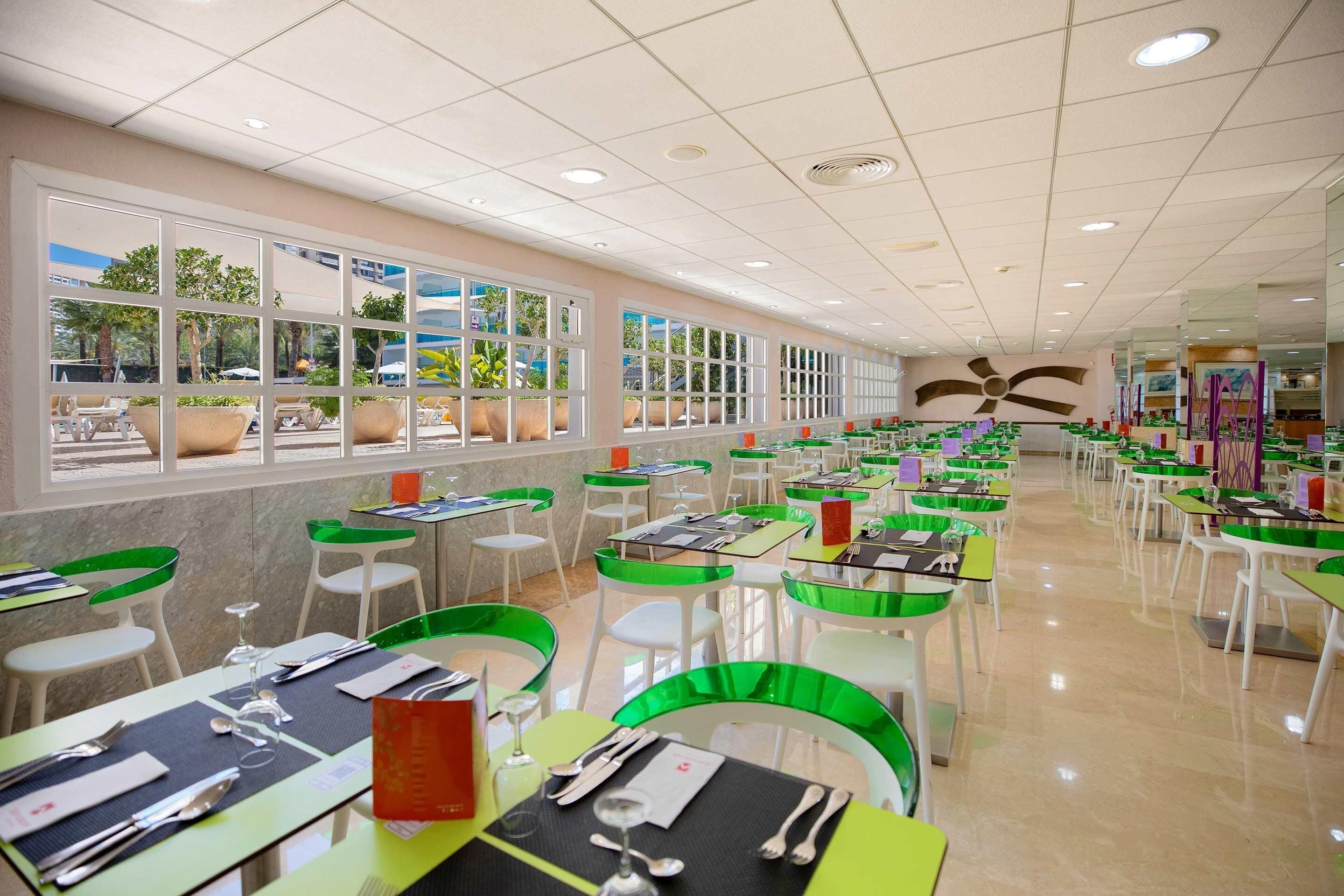 A spacious dining area with multiple tables and chairs, large windows with views of the outdoors, and a ceiling fan.