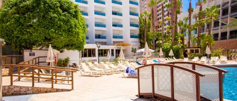 A hotel with a swimming pool, lounge chairs, and a large building with balconies.