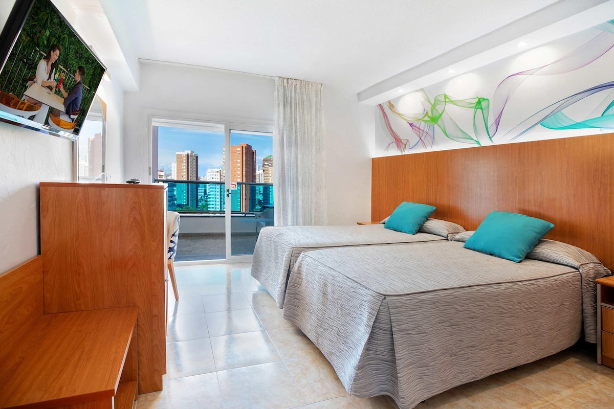 Ein Hotelzimmer mit Balkon, einem Bett mit zwei Kissen, einem Holztisch und einem an der Wand befestigten Fernseher.