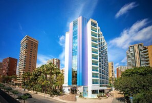 A modern high-rise hotel with a prominent vertical blue and white facade, surrounded by palm trees and other buildings. - Hotel Servigroup Castilla (Benidorm)