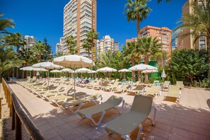 A resort pool area with sun loungers and umbrellas, surrounded by palm trees and tall buildings. - Hotel Servigroup Castilla (Benidorm)