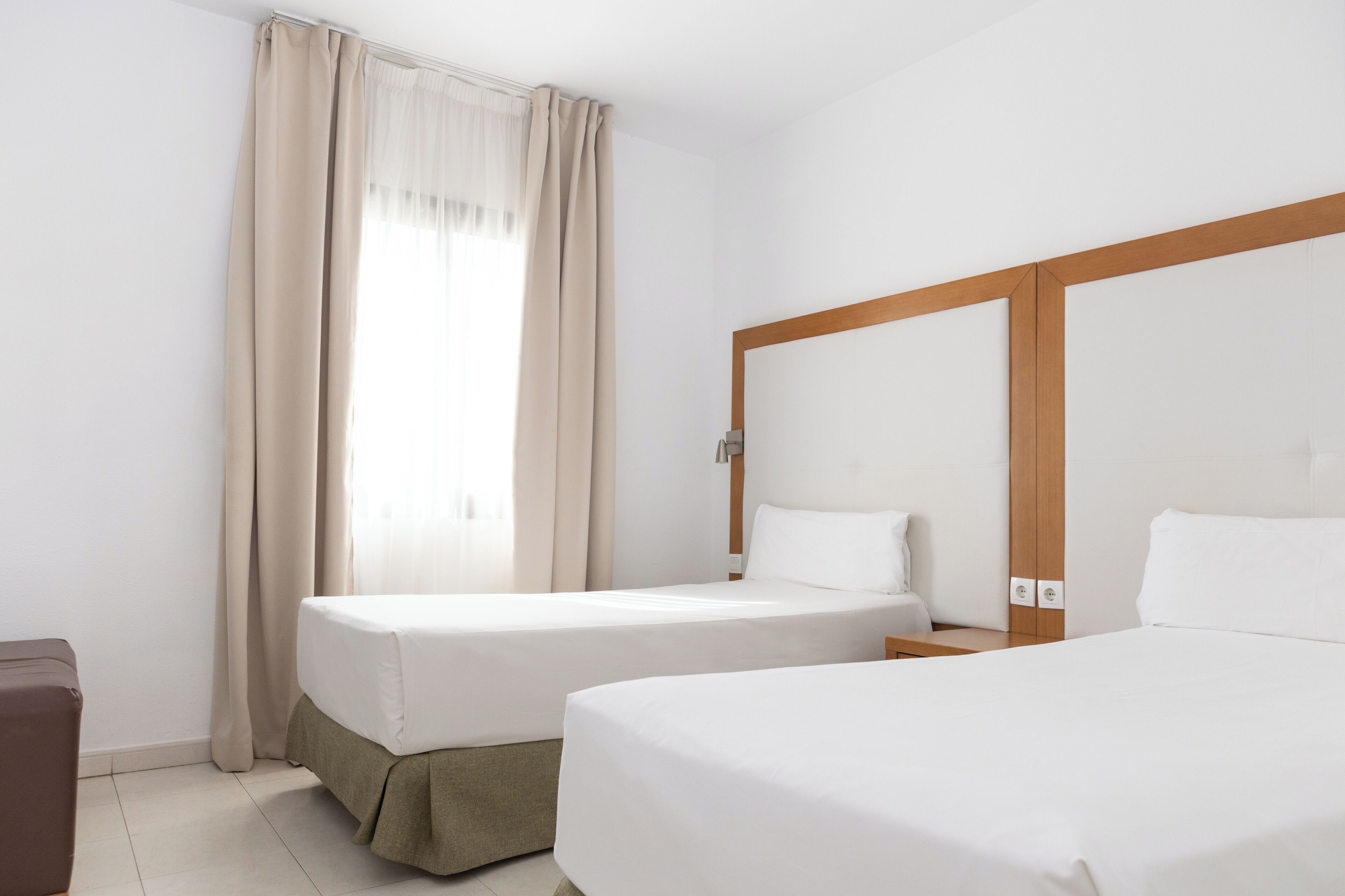 A hotel room with two beds, a wooden headboard, a window with curtains, and a small bedside table.