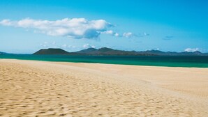 A beach with clear turquoise water and distant mountains under a blue sky. - Apartamentos Hesperia Bristol Playa (La Oliva)