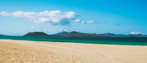 A beach with clear turquoise water and distant mountains under a blue sky.