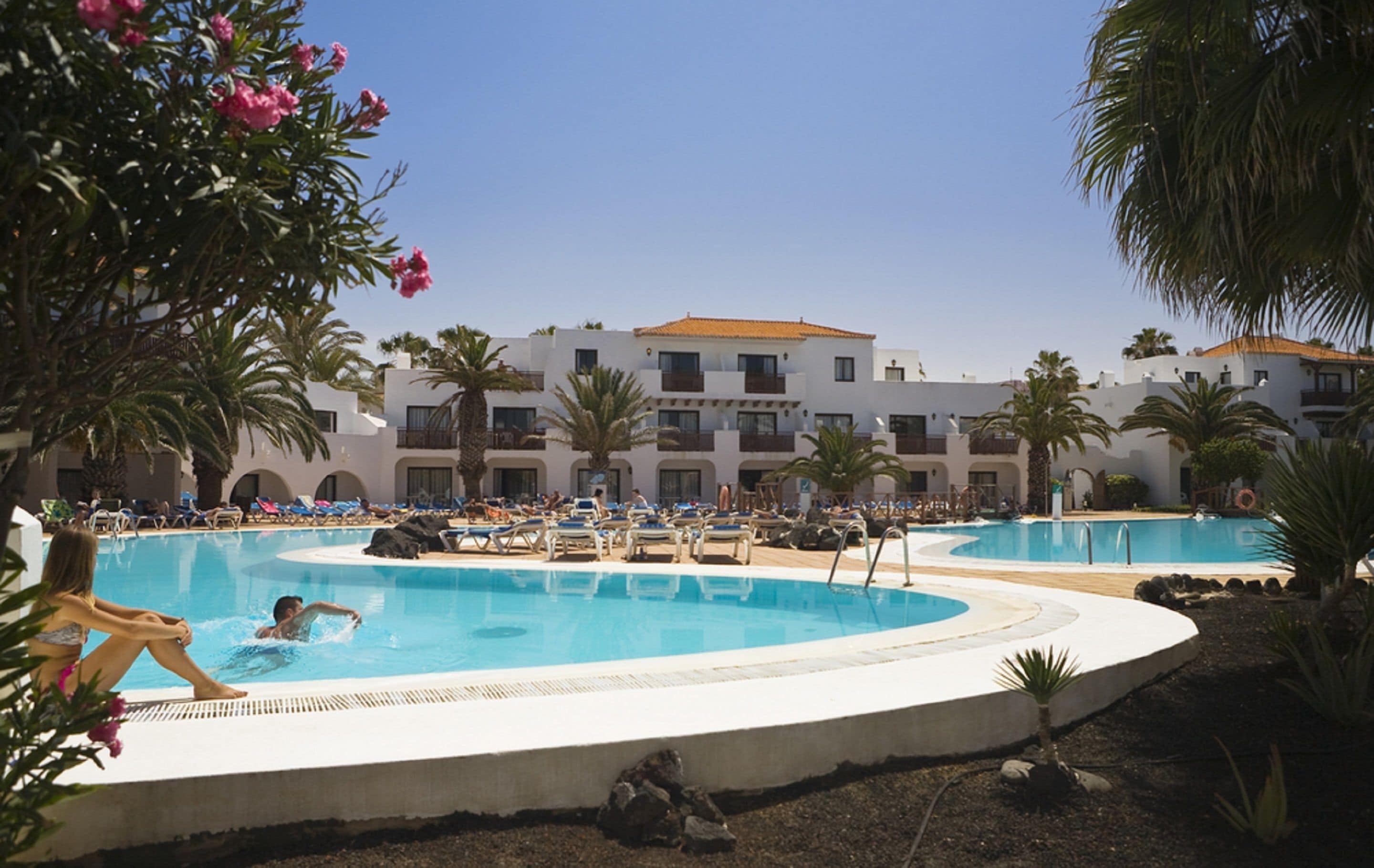 Un área de piscina en un resort, con varias piscinas, palmeras y un edificio con balcones al fondo.
