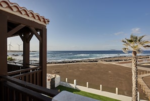 A beach view from a covered area with a wooden deck, a palm tree, and wind turbines in the background.