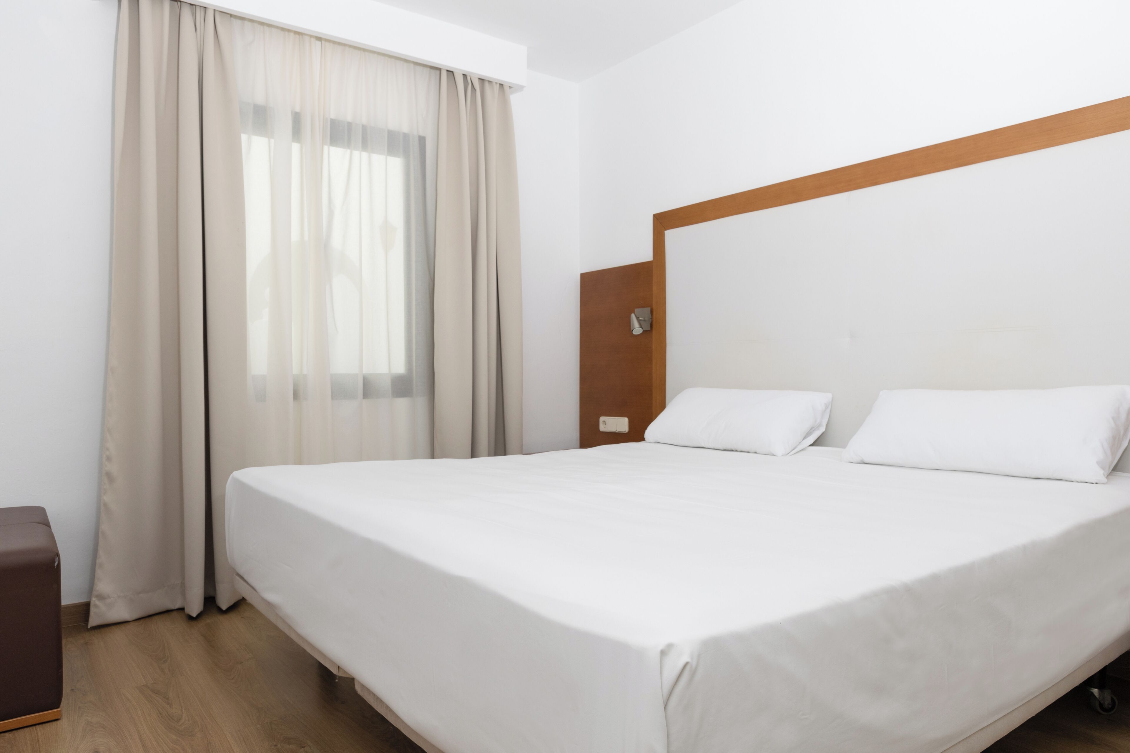 A hotel room with a large bed, a wooden headboard, a small bedside table, and a window with curtains.