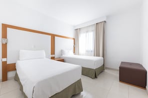 Habitación de hotel con dos camas, cabecera de madera, una otomana marrón y una ventana con cortinas.