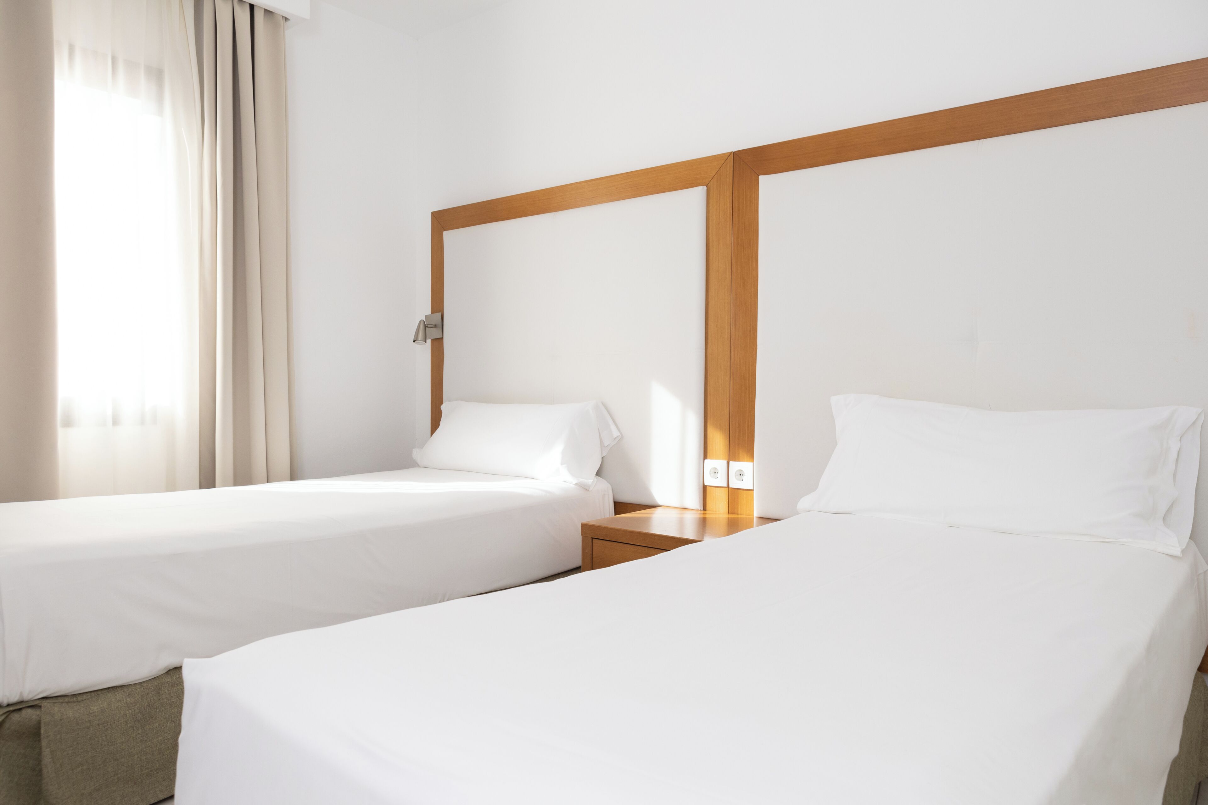 Une chambre d’hôtel avec deux lits simples, une tête de lit en bois, une table de chevet, une lampe et une fenêtre avec des rideaux.