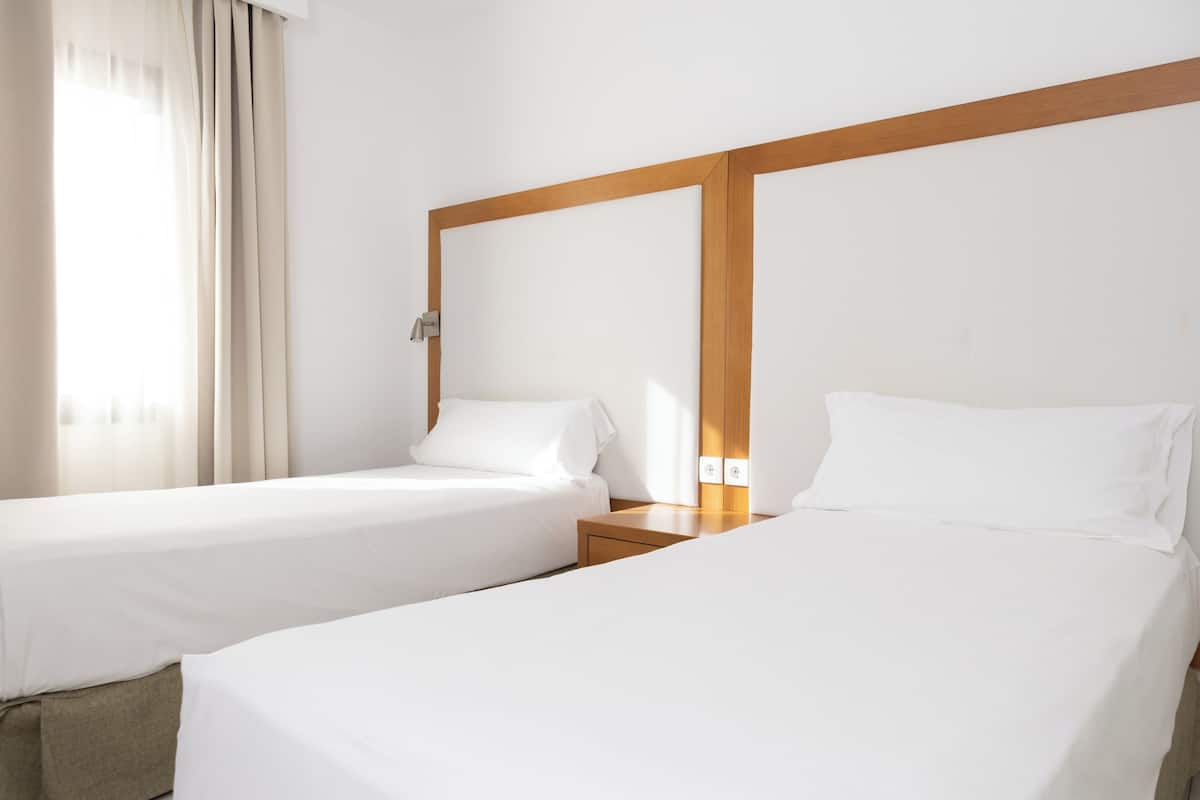 Una habitación de hotel con dos camas individuales, cabecera de madera, mesita de noche, lámpara y ventana con cortinas.