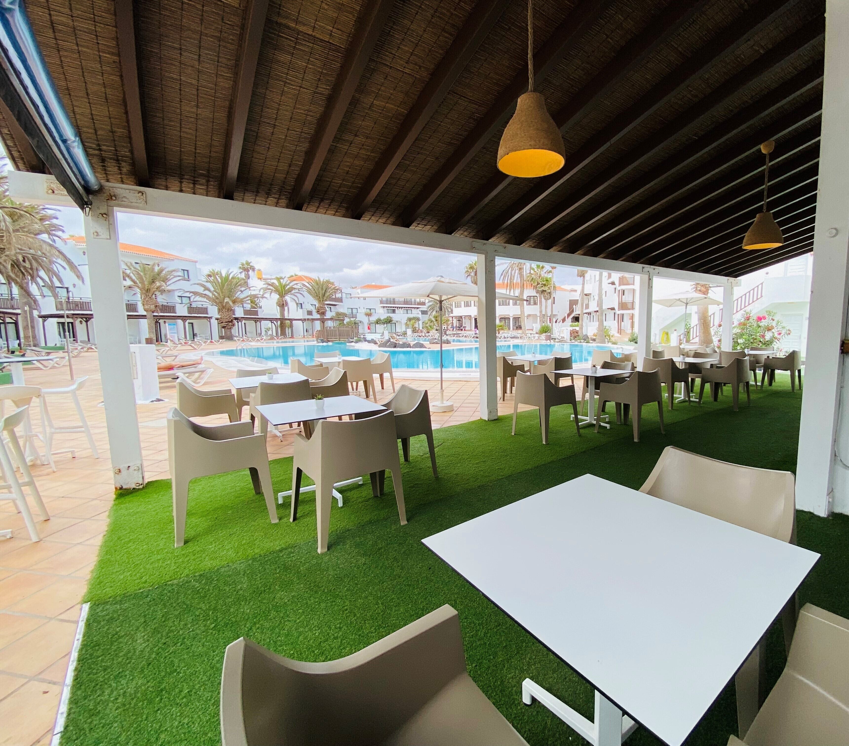 Un espacio al aire libre con mesas y sillas, piscina y vistas a los edificios del complejo turístico.