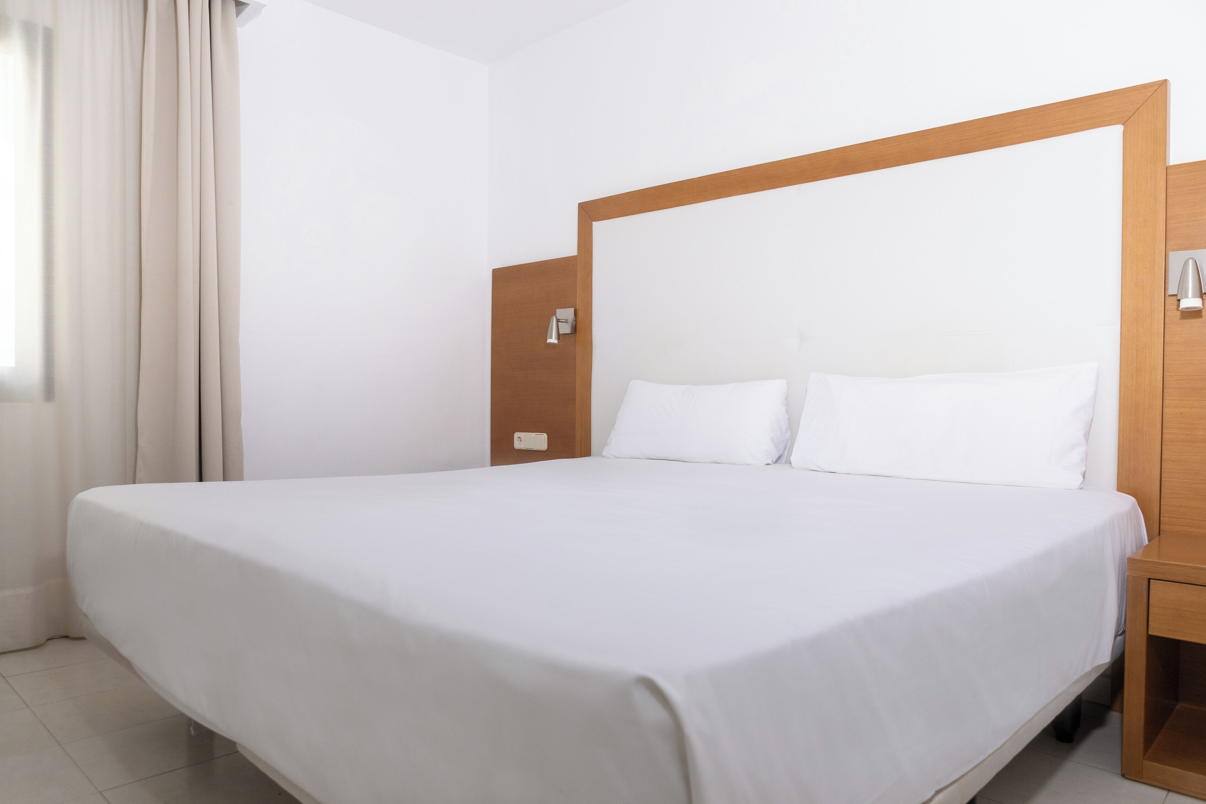 A neatly made bed in a hotel room with a wooden headboard, a bedside table, and a wall-mounted light.