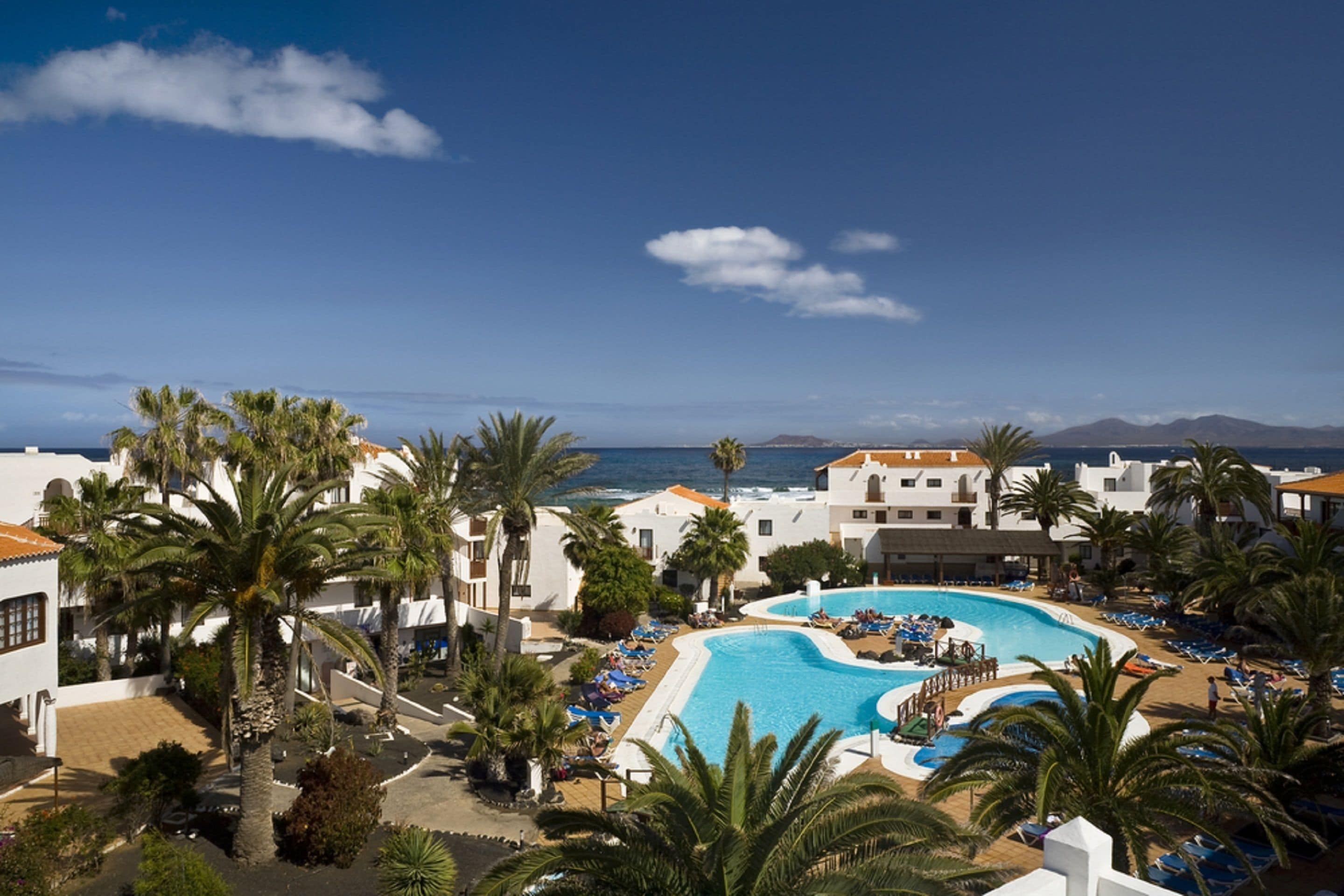 Un resort con una gran piscina rodeada de palmeras y edificios blancos, con vistas al mar.