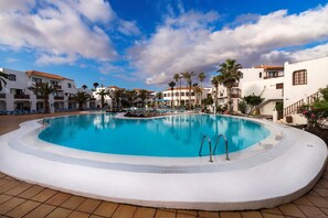 A large, clear swimming pool surrounded by white buildings with balconies, palm trees, and a blue sky with clouds.