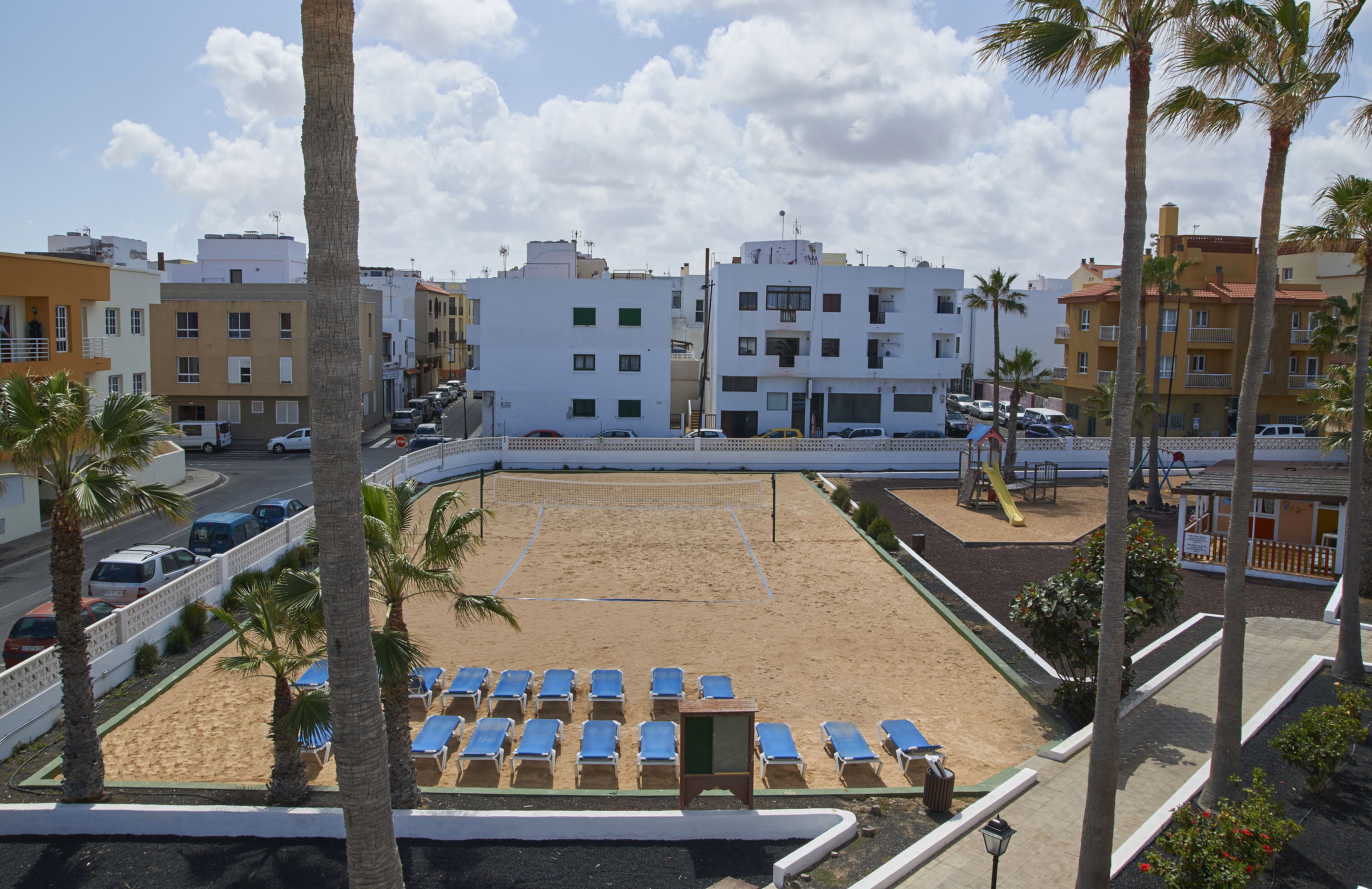 A beach with blue lounge chairs and a volleyball net, surrounded by palm trees and buildings.
