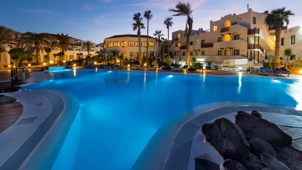 A large swimming pool with lounge chairs and palm trees, surrounded by buildings with balconies and warm lighting. - Apartamentos Hesperia Bristol Playa (La Oliva)