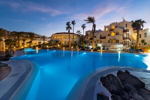 A large swimming pool with lounge chairs and palm trees, surrounded by buildings with balconies and warm lighting. - Apartamentos Hesperia Bristol Playa (La Oliva)