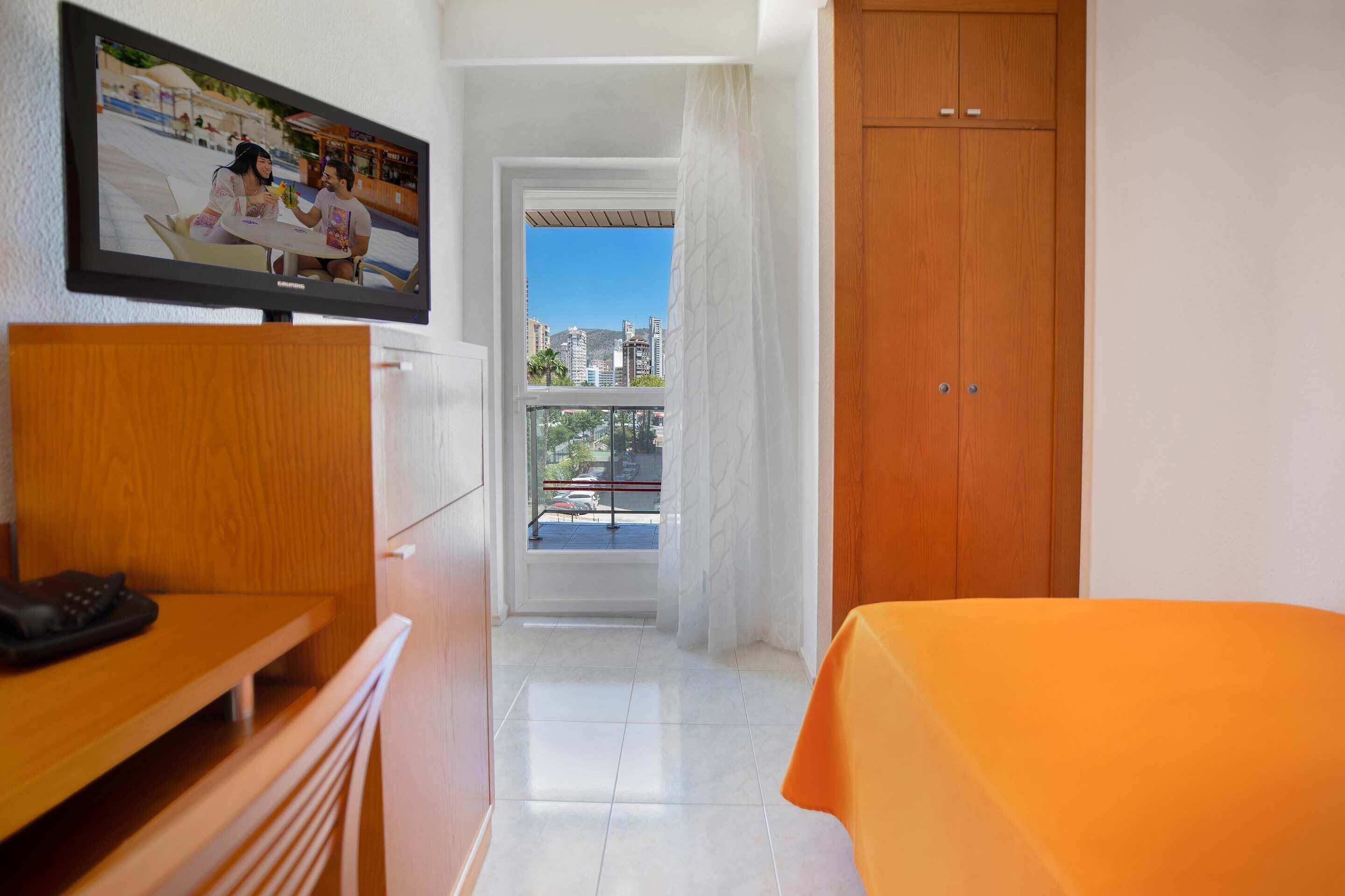A hotel room with a bed, a TV mounted on the wall, a desk, and a view of the city through a window.