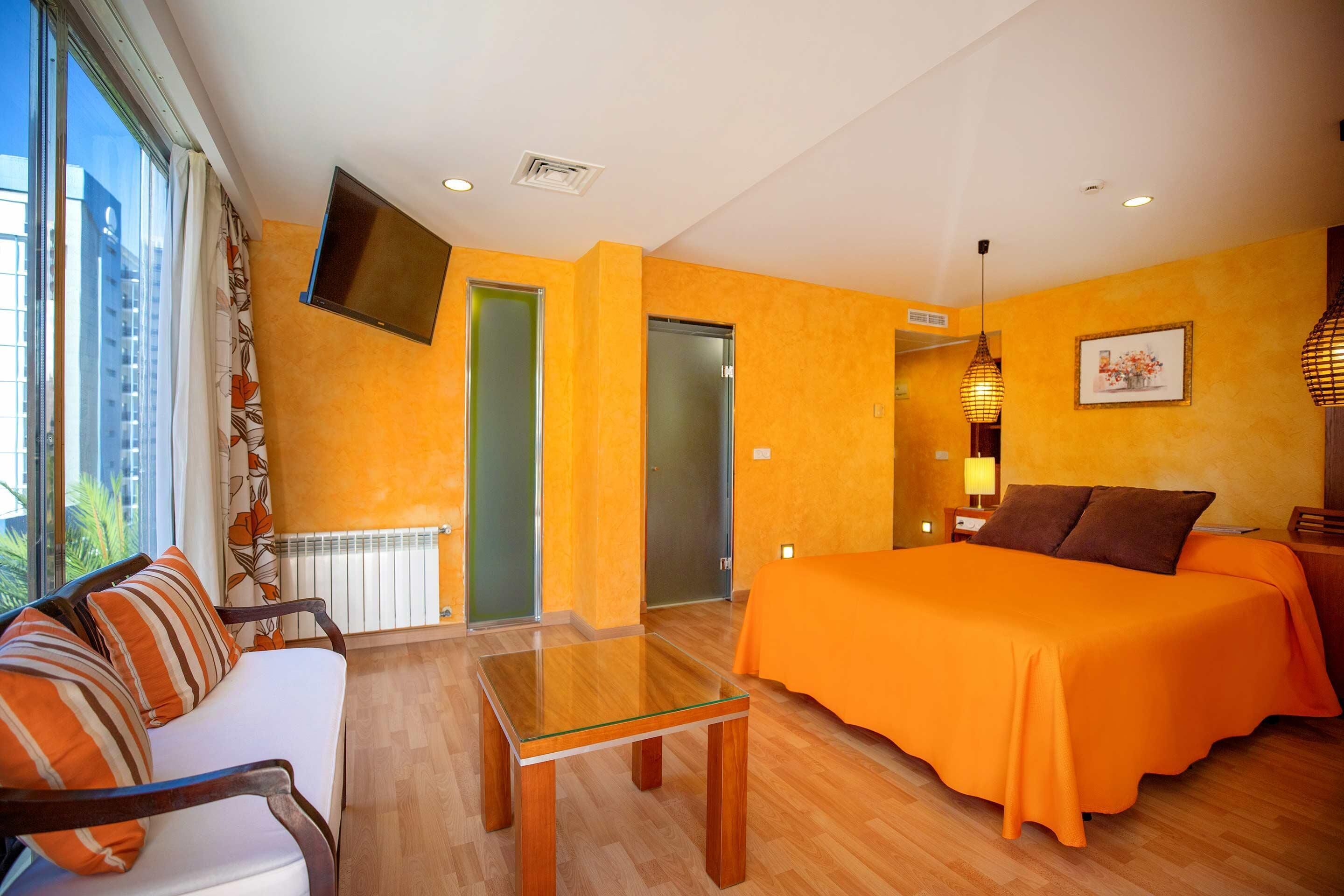 A hotel room with a bed, a sofa, a glass table, and a TV mounted on the wall.