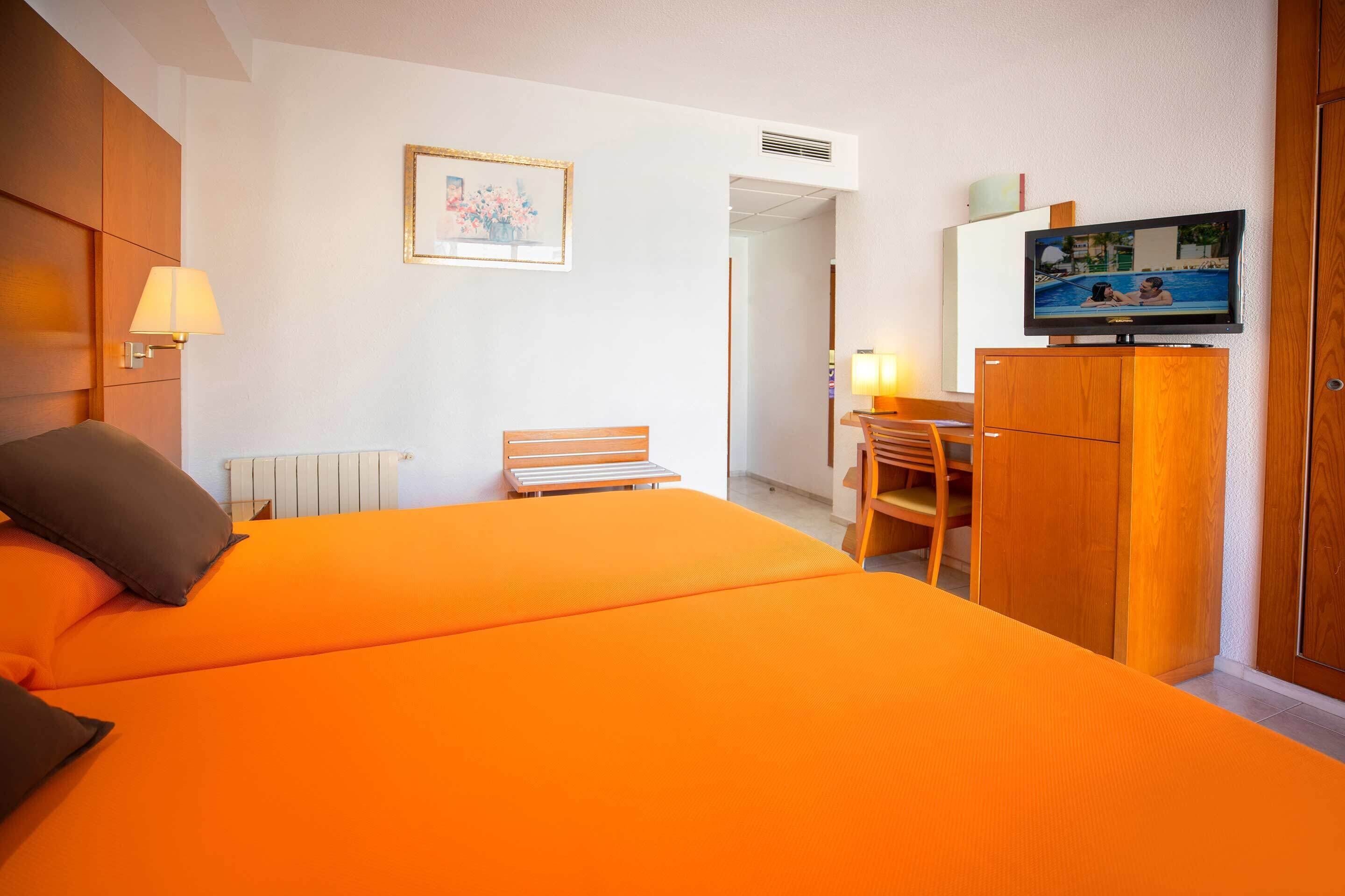A hotel room with a large orange bed, a desk with a chair, a television on a wooden cabinet, and a framed picture on the wall.