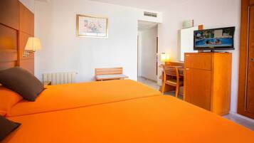 Habitación de hotel con una cama grande color naranja, un escritorio con silla, un televisor sobre un mueble de madera y un cuadro en la pared.