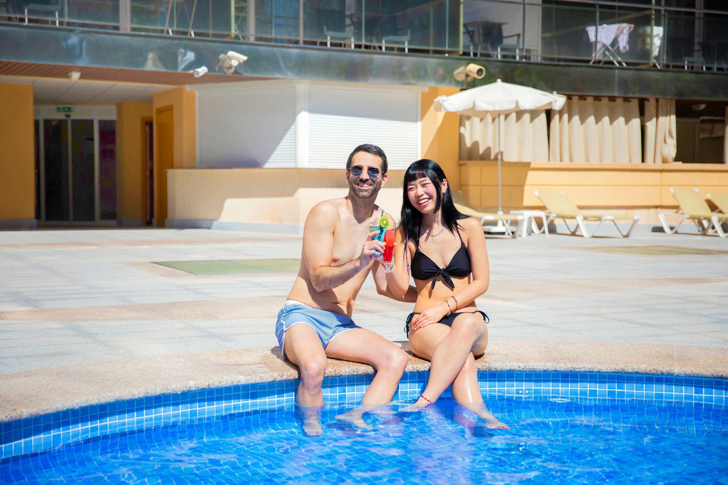 A couple sitting by a swimming pool with lounge chairs and umbrellas in the background.