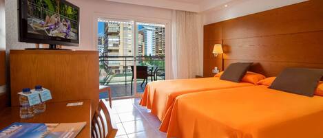 Habitación de hotel con dos camas, televisión, escritorio con silla y balcón con vistas a edificios.