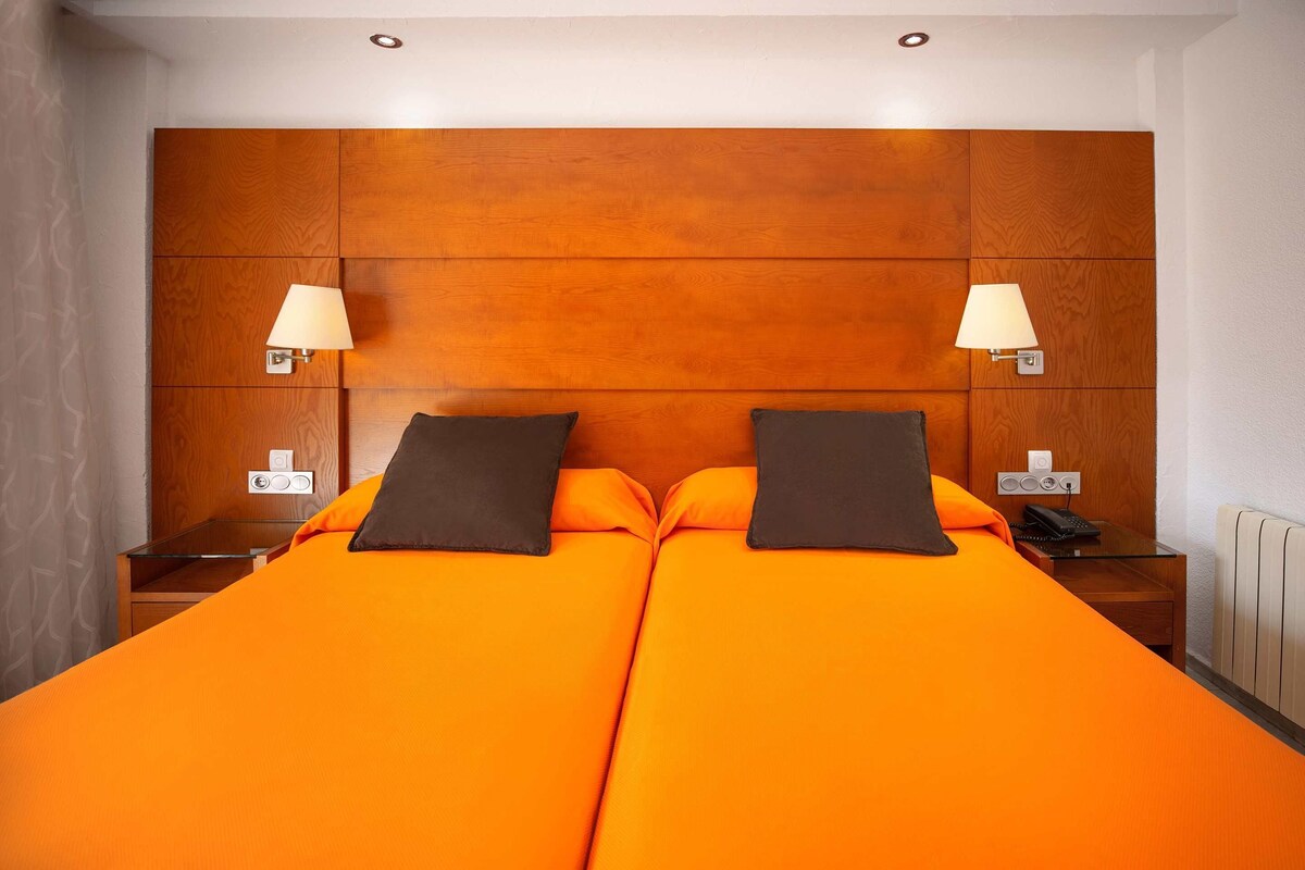 Una cama doble con ropa de cama naranja y dos almohadas oscuras, flanqueada por mesitas de noche con lámparas y un teléfono.