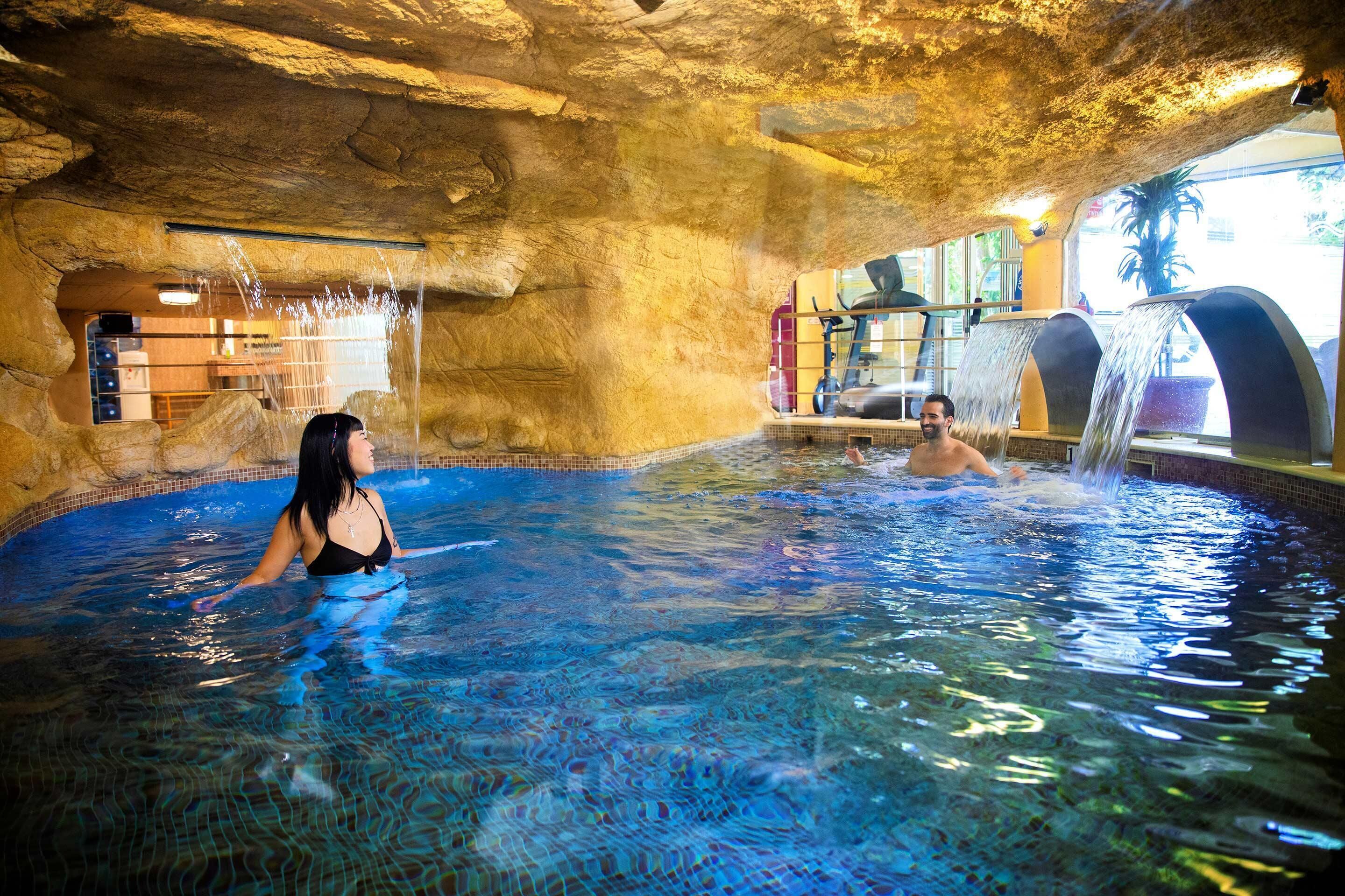 An indoor pool with clear blue water, a rocky cave-like ceiling, and a waterfall feature.