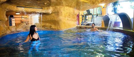 An indoor pool with clear blue water, a rocky cave-like ceiling, and a waterfall feature.