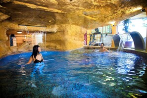 Une piscine intérieure à l'eau d'un bleu cristallin, avec un plafond rappelant une grotte rocheuse et une cascade décorative.